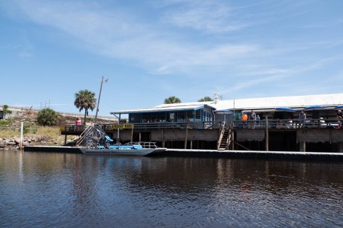 A waterfront restaurant with a boat dock, surrounded by palm trees and a clear blue sky.