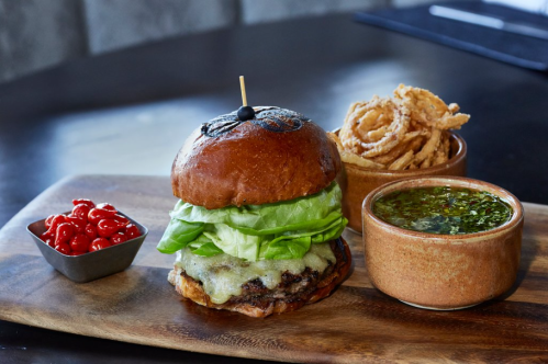 A gourmet burger with melted cheese, lettuce, and a skewer, served with onion rings and a side of sauce and cherry tomatoes.