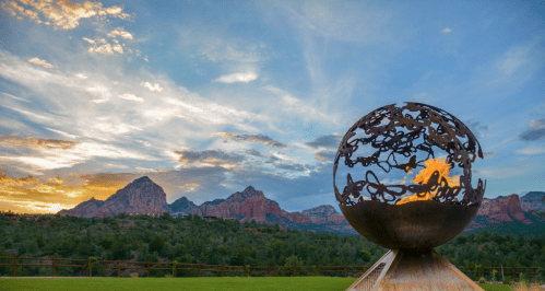 A metal globe sculpture with flames, set against a sunset over red rock mountains and a cloudy sky.