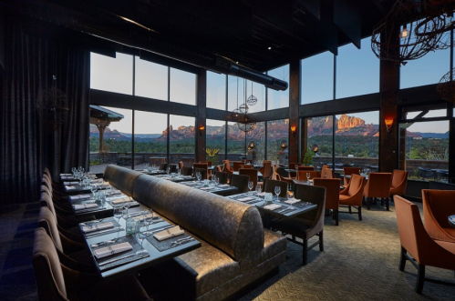 Elegant restaurant interior with a long table, plush seating, and large windows showcasing a mountain view at sunset.