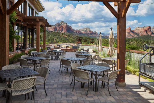 Outdoor patio with wicker tables and chairs, overlooking scenic mountains under a partly cloudy sky.