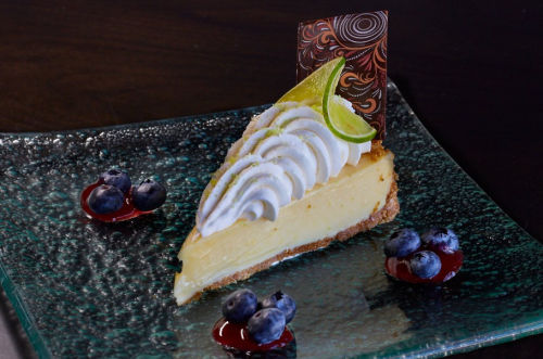 A slice of cheesecake topped with whipped cream, lime, and chocolate, garnished with blueberries and berry sauce on a glass plate.