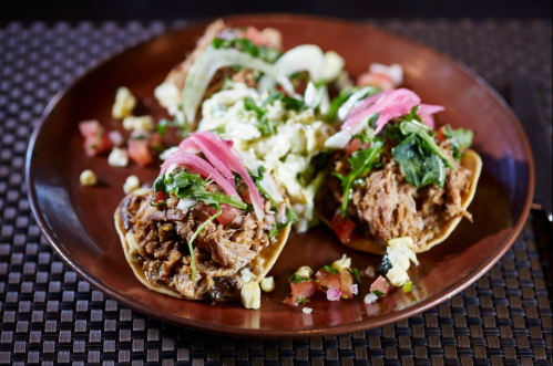 A plate of three tacos topped with onions, pickled vegetables, and fresh herbs, served with a side of salad and salsa.