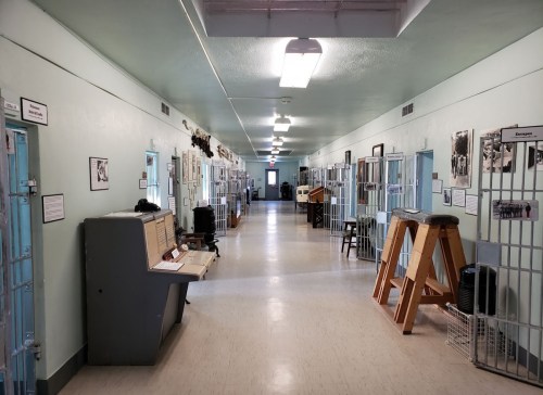 A long, narrow hallway in a prison with blue cell doors, historical photos, and display cases along the walls.