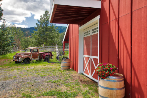 A rustic red barn with a vintage truck parked nearby, surrounded by greenery and flower-filled barrels.
