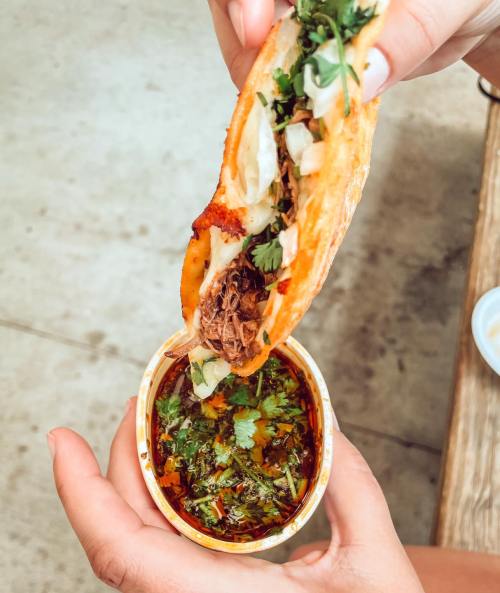 A hand holding a taco filled with meat and toppings, above a bowl of vibrant green salsa with herbs.