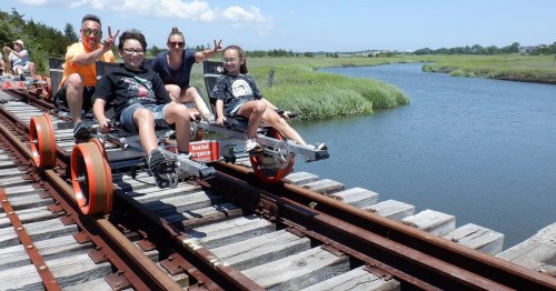 A group of four people riding a rail bike on a wooden track beside a river, surrounded by greenery and blue skies.