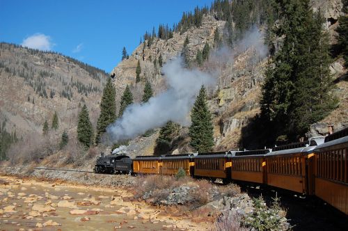 A steam train travels along a scenic mountain landscape, with smoke billowing and trees lining the tracks.
