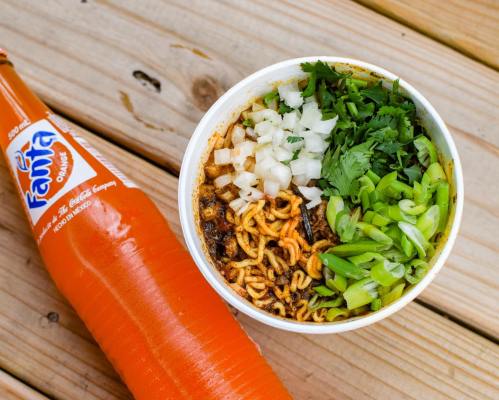 A bowl of spicy noodles topped with chopped onions and green onions, alongside a bottle of orange Fanta.