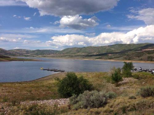 A serene lake surrounded by green hills and blue skies with fluffy clouds, featuring a small dock and boats.