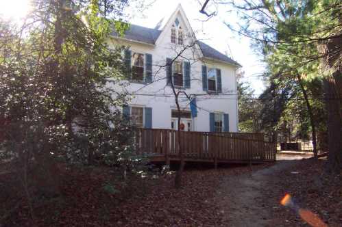 A white two-story house surrounded by trees, with a wooden deck and a path leading up to it.