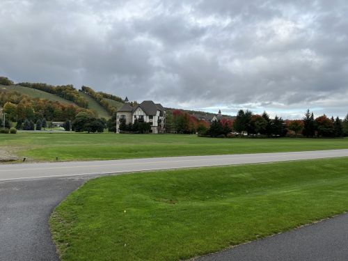 A large building surrounded by green grass and trees, with a hillside in the background displaying autumn colors under a cloudy sky.