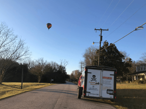 A hot air balloon floats above a quiet street with a parked trailer on the side, surrounded by trees and clear skies.
