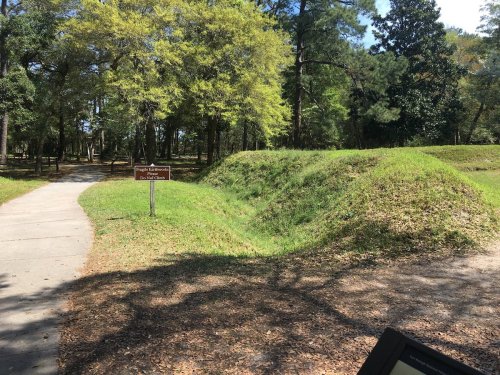 A grassy area with a sign indicating a historical site, surrounded by trees and a walking path.