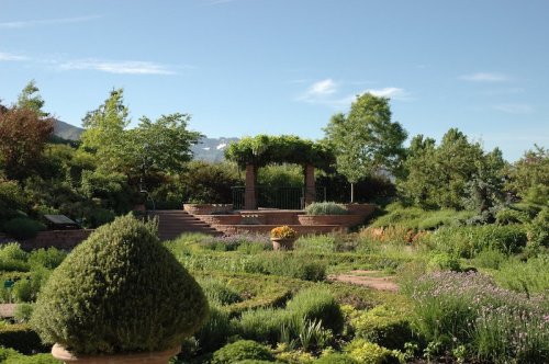 A lush garden with a stone gazebo, surrounded by greenery and colorful flowers under a clear blue sky.