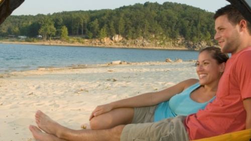 A couple relaxes on a beach, smiling and enjoying the view of the water and trees in the background.
