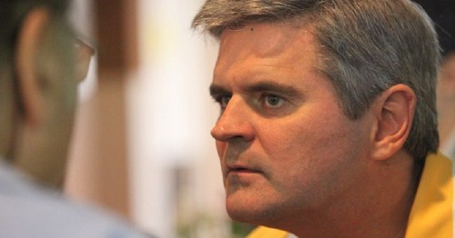 A man with gray hair looks intently at another person, engaged in a serious conversation.