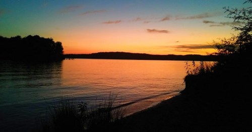 A serene sunset over a calm lake, with silhouettes of trees and gentle waves along the shore.