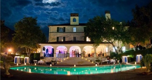 A beautifully lit mansion at night, with a pool in the foreground and guests enjoying the outdoor space.