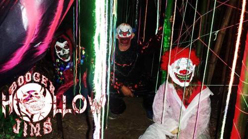 Three clowns with eerie makeup sit in a dark, colorful setting, surrounded by glowing lights and spooky decorations.