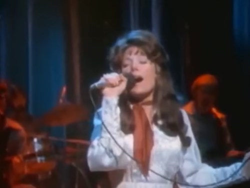 A woman sings passionately into a microphone on stage, with a band playing in the background.