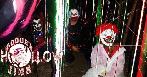 Three clowns with spooky makeup pose in a dark, colorful setting filled with lights and decorations.