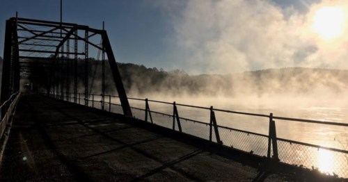 A misty morning scene of a bridge over a river, with fog rising and sunlight illuminating the landscape.