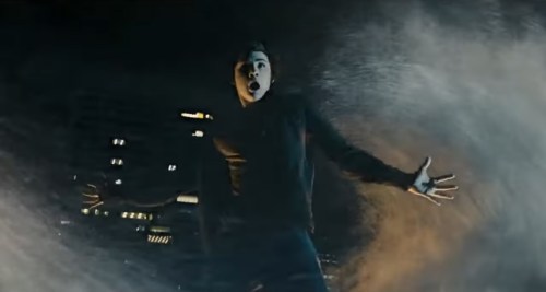 A person stands with arms outstretched, looking surprised amidst swirling mist or smoke at night.