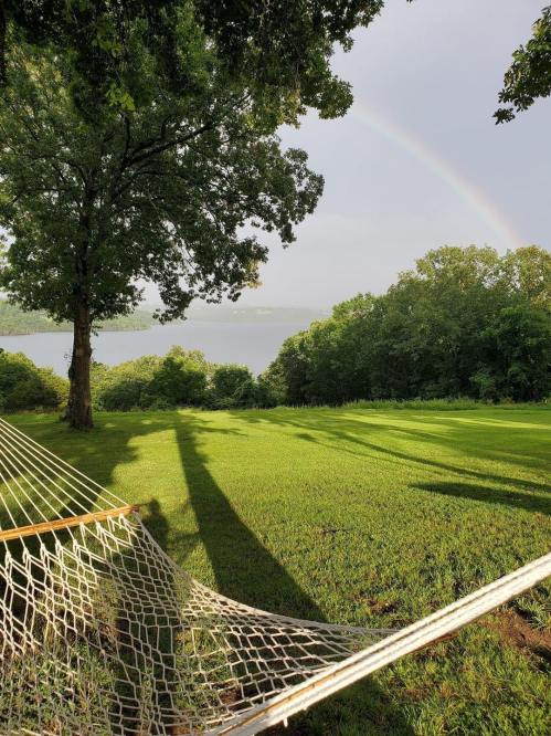 A hammock under a tree, with a lush green lawn and a rainbow in the background over a serene lake.