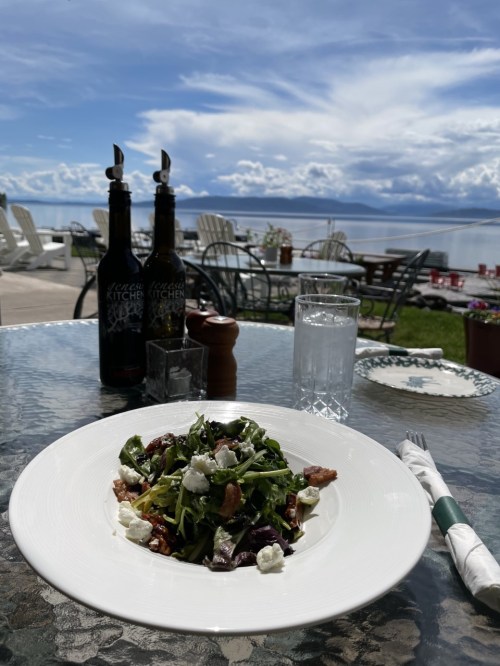 A fresh salad with greens and goat cheese on a table by the water, with bottles and glasses in the background.