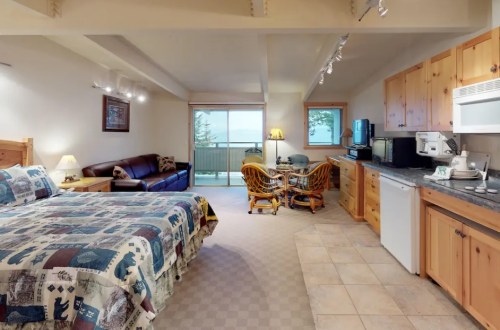 Cozy hotel room with a bed, seating area, kitchenette, and large window overlooking a balcony. Warm wooden decor.