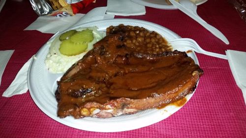 A plate of barbecue ribs with sauce, coleslaw, pickles, and baked beans on a white disposable plate.