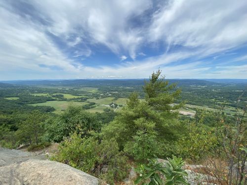 A panoramic view from a mountain top, showcasing lush green valleys and a blue sky with scattered clouds.