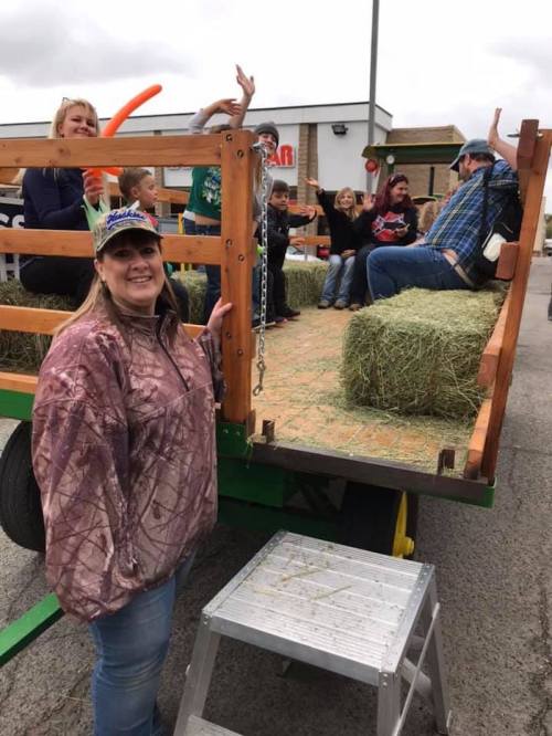 A woman in a camouflage jacket stands by a hay-filled wagon with children and adults enjoying a festive event.