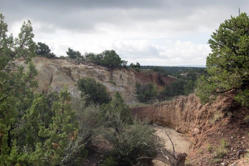 A rugged landscape featuring cliffs, greenery, and a dry riverbed under a cloudy sky.