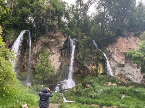 A person stands in front of cascading waterfalls surrounded by lush greenery and rocky cliffs.