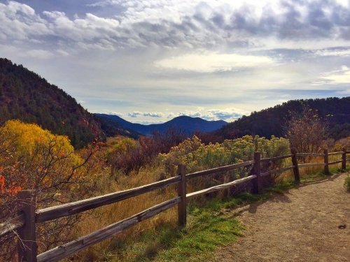 Scenic view of mountains and valleys with colorful foliage, framed by a wooden fence under a cloudy sky.