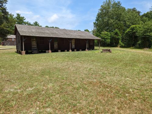 A rustic wooden building with a sloped roof, set in a grassy area surrounded by trees under a blue sky.