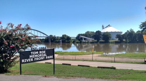 Sign for Tom Roe Memorial River Front Park with a view of the river and a bridge in the background on a clear day.