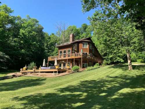 A large wooden house surrounded by lush greenery and trees, with a deck and patio area in a sunny outdoor setting.