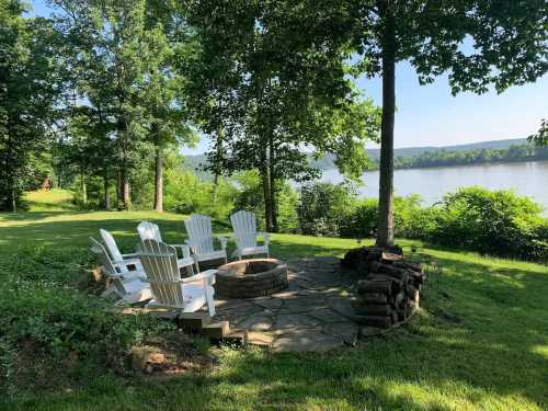 A serene lakeside scene featuring a stone fire pit surrounded by white chairs and lush greenery.