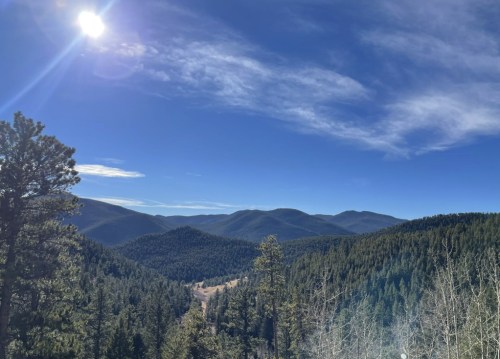 A scenic view of mountains and forests under a clear blue sky, with the sun shining brightly in the corner.