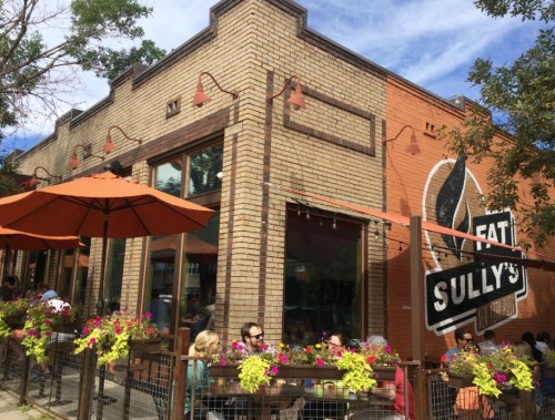A brick restaurant with outdoor seating, colorful flowers, and an orange umbrella, featuring a sign that reads "Eat Sully's."