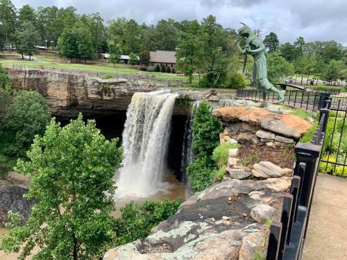 A statue of a figure stands near a waterfall, surrounded by lush greenery and a scenic landscape.