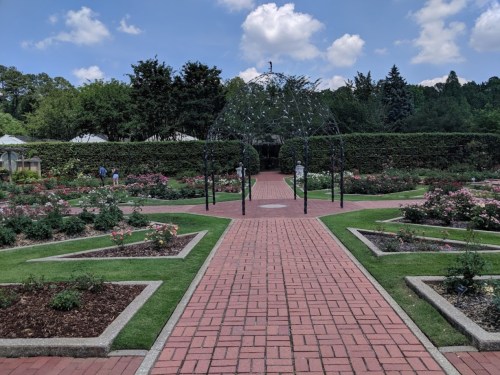 A landscaped garden with a brick pathway, rose bushes, and a decorative archway under a blue sky with clouds.