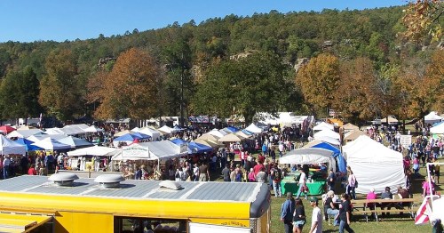 A bustling outdoor market with numerous tents and crowds, surrounded by trees with autumn foliage.