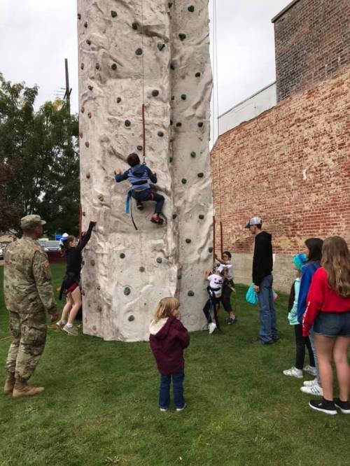 A group of children and adults watch as a child climbs a rock wall, with a soldier standing nearby.