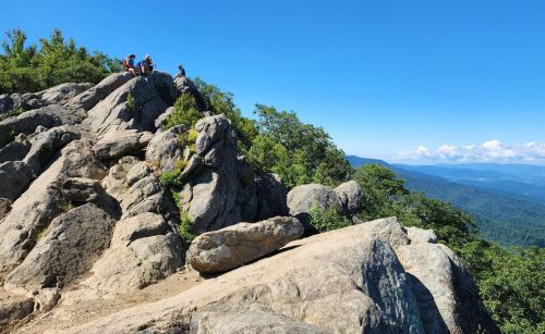 A rocky mountain peak with hikers enjoying the view under a clear blue sky and lush green surroundings.