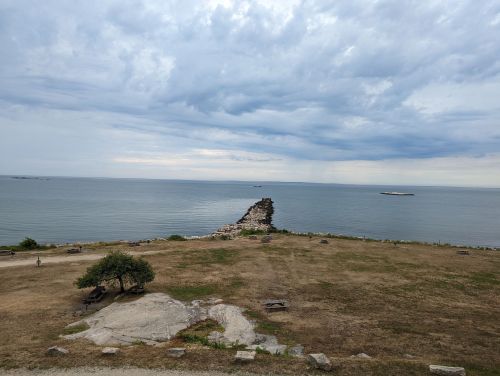 A rocky shoreline extends into a calm sea under a cloudy sky, with sparse grass and a few trees in the foreground.