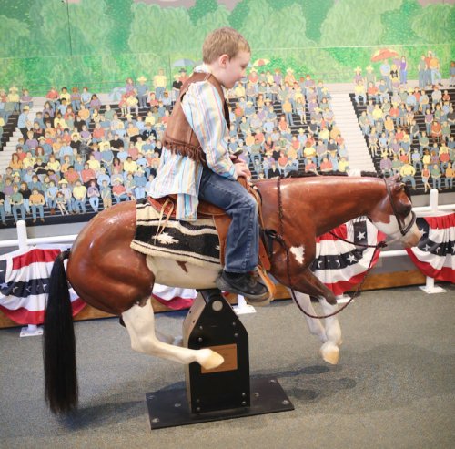 A young boy in a cowboy outfit rides a mechanical horse in front of a colorful crowd mural.
